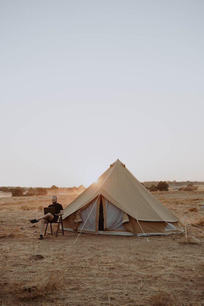 Full body of male traveler sitting on chair with laptop and working during vacation in camping