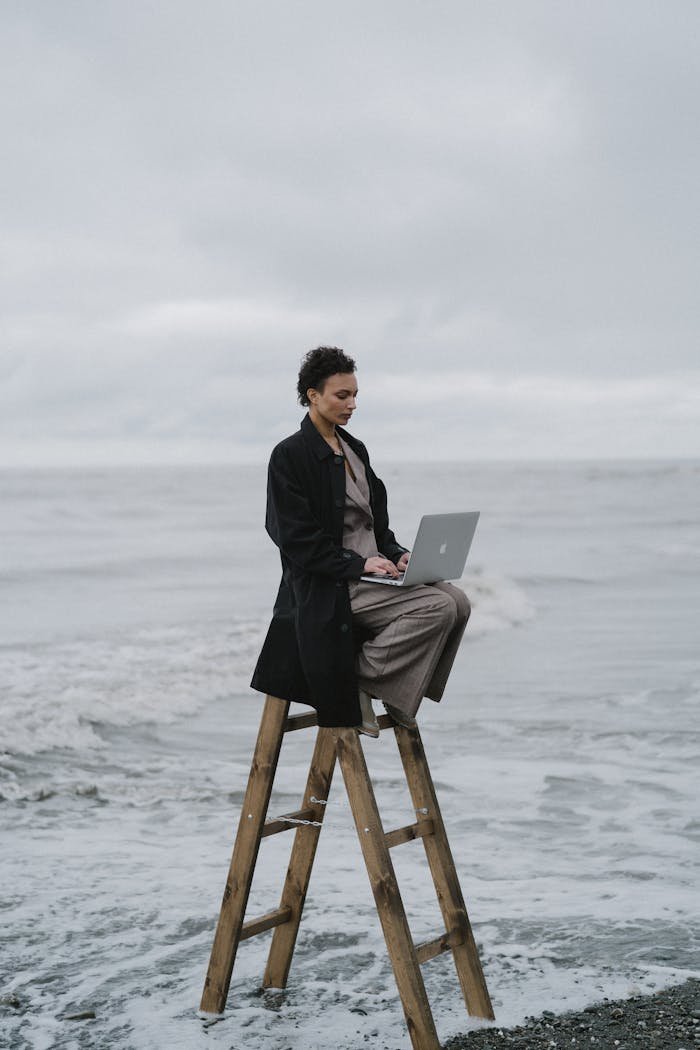 A Woman Sitting on a Wooden Ladder at the Beach while Using Her Laptop